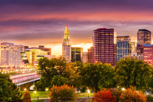 Connecticut - Shutterstock | CareersinPsychology.org A vibrant cityscape of Hartford, Connecticut, featuring a skyline with modern skyscrapers and the distinctive Travelers Tower at sunset, surrounded by lush green trees and a river, symbolizing the urban and natural beauty of the state where social work opportunities abound.