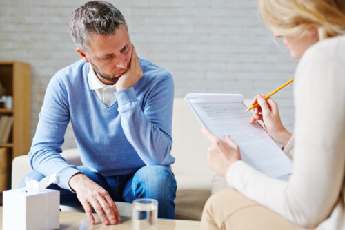 psychiatrist conducting therapy session with male patient in modern clinical office, counselor taking notes during mental health consultation